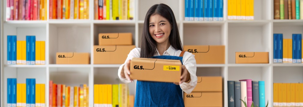 Woman handing out a GLS parcel. She stands at a desk in front of shelves field with colourful books.